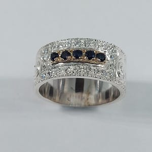May include: A silver ring with a wide band, encrusted with small, clear stones. A central section features five dark blue gemstones set in a gold-colored band. The ring has decorative scrollwork on the sides, creating an elegant and detailed design.