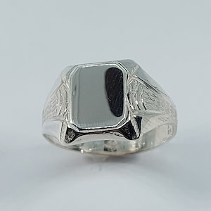 Puede incluir: Un anillo de sello de plata con una parte superior rectangular lisa y detalles grabados intrincados en la banda.