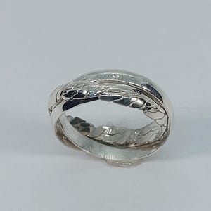 May include: A silver ring with a textured, interwoven design. The ring features overlapping bands, creating a three-dimensional effect. The surface of the ring has a detailed pattern, adding to its intricate appearance. The ring is likely made of sterling silver.