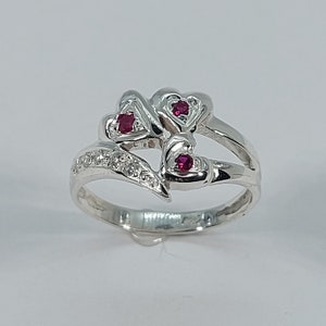 Könnte beinhalten: Ein silberner Ring mit einem filigranen Design, das drei rote Edelsteine in Herzform zeigt. Der Ring ist mit kleinen weißen Edelsteinen verziert.