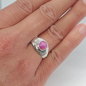 K&ouml;nnte beinhalten: Ein silberner Ring mit einem rosafarbenen Edelstein, der in einer runden Fassung sitzt. Der Ring hat eine strukturierte Band mit mehreren Rillen.
