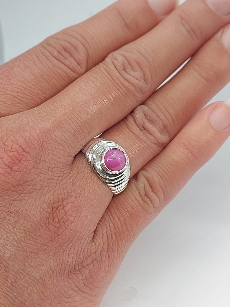 K&ouml;nnte beinhalten: Ein silberner Ring mit einem rosafarbenen Edelstein, der in einer runden Fassung sitzt. Der Ring hat eine strukturierte Band mit mehreren Rillen.