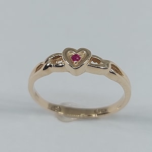 Peut inclure: Une bague dorée avec un cœur serti d'une petite pierre rouge ronde. La monture présente un motif délicat et entrelacé. La bague est présentée sur un fond clair et uni.