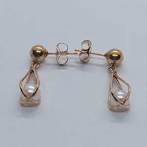 May include: A pair of gold earrings with a pearl drop. The earrings have a simple design with a small gold ball at the top and a delicate cage holding a single pearl.