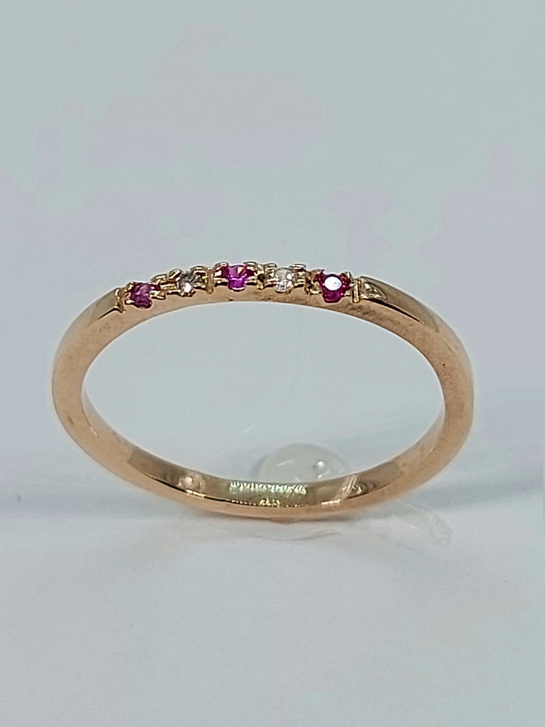 Pode incluir: Um delicado anel de banda de ouro com uma fileira de pequenas pedras preciosas rosa e brancas.