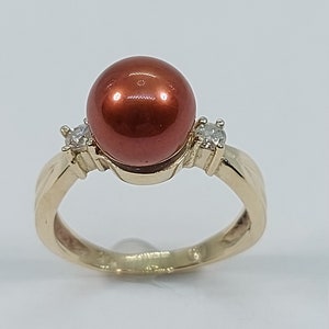 Può includere: Un anello d'oro con una grande perla rotonda rossa e due piccoli diamanti rotondi trasparenti.