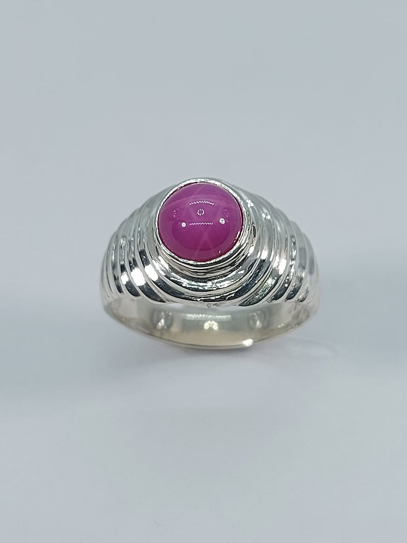 K&ouml;nnte beinhalten: Ein silberner Ring mit einem rosafarbenen Cabochon-Edelstein, der in einer erh&ouml;hten Fassung sitzt. Die Ringschiene hat ein strukturiertes, geriffeltes Design.