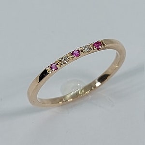 Pode incluir: Um delicado anel de banda de ouro com uma fileira de pequenas pedras preciosas rosa e brancas.