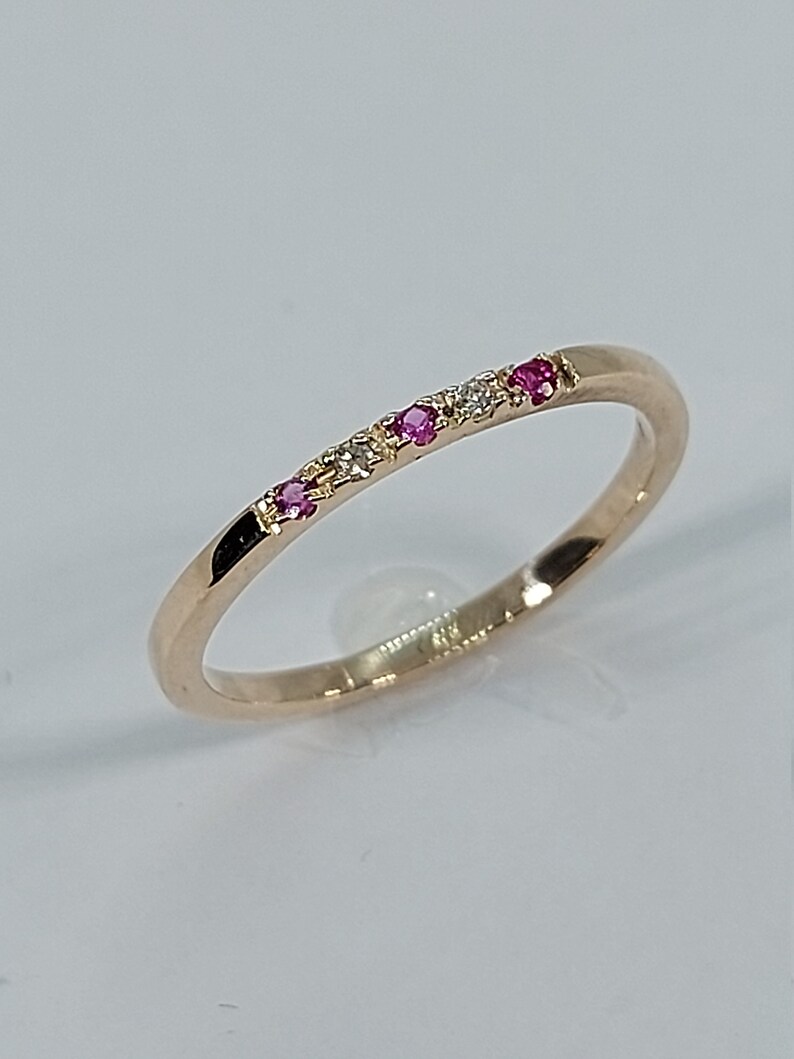 Pode incluir: Um delicado anel de banda de ouro com uma fileira de pequenas pedras preciosas rosa e brancas.