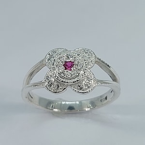 May include: A silver ring featuring a four-leaf clover design. The centre of the clover is a red gemstone surrounded by smaller white gemstones.