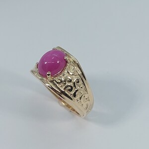 Op de afbeelding: Een gouden ring met een ovale roze edelsteen in het midden. De band van de ring is versierd met een ingewikkeld bloemmotief.
