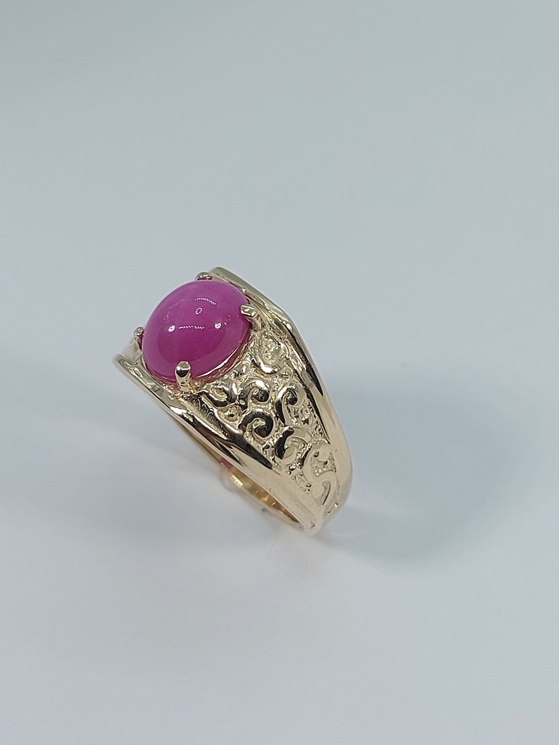Op de afbeelding: Een gouden ring met een ovale roze edelsteen in het midden. De band van de ring is versierd met een ingewikkeld bloemmotief.