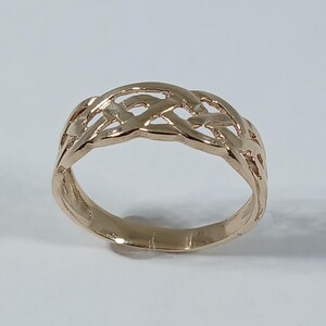 May include: A gold-colored ring with a Celtic knot design. The ring features an openwork pattern of interwoven strands, creating a continuous, intricate design. The ring is set against a plain white background.