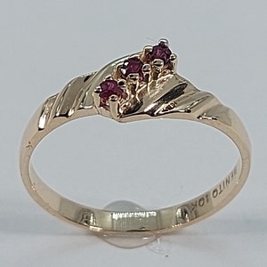 以下が含まれることがあります： ねじれたバンドに3つの小さなルビーレッドの石がセットされたゴールドリング。リングには「BENITO JOY」という文字が刻まれています。