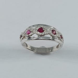May include: A silver ring with a series of small, round, red gemstones set in a decorative pattern. The ring has a polished finish and a classic design, suitable for everyday wear or special occasions.