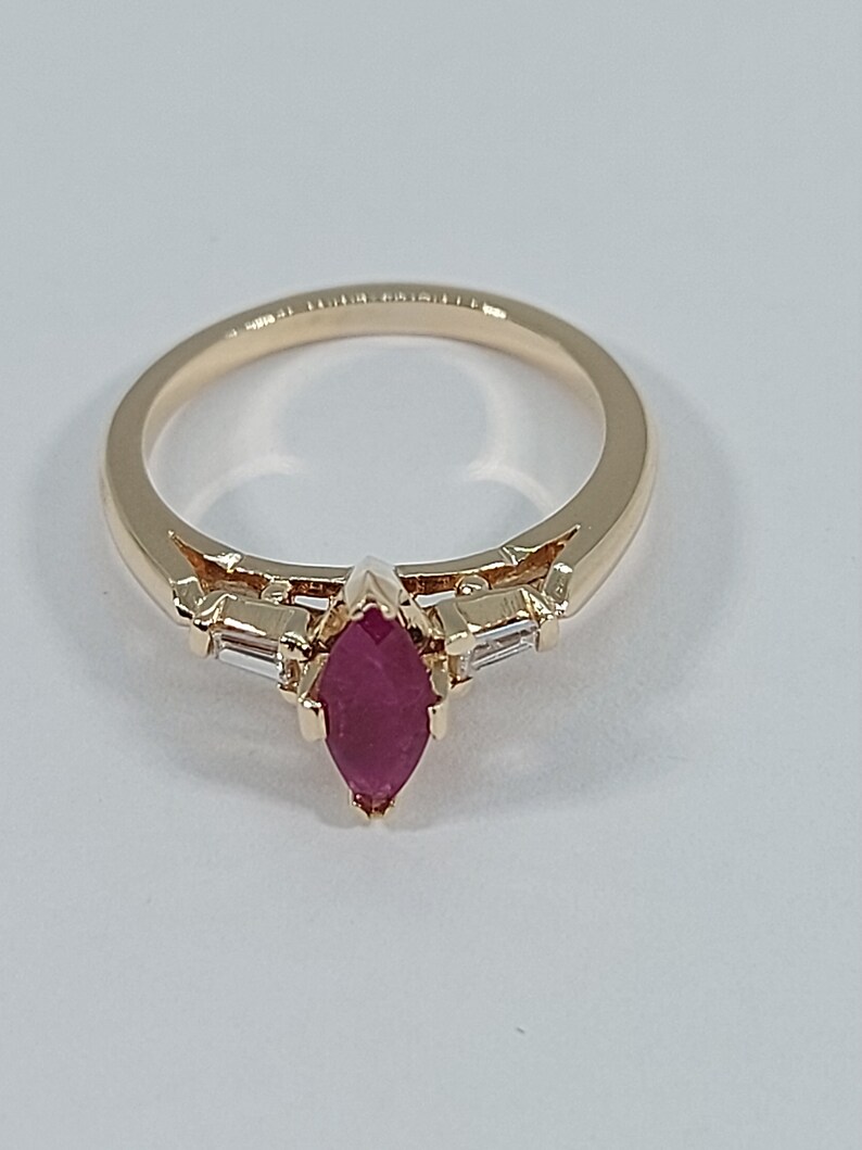 May include: A gold ring featuring a marquise-shaped ruby gemstone, flanked by two smaller baguette-shaped diamonds.