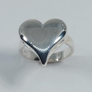 May include: A polished silver ring featuring a large, heart-shaped design. The heart has a smooth, reflective surface and is mounted on a simple band. The ring is a close-up shot against a plain background.
