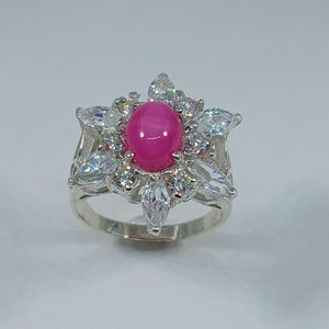 Op de afbeelding: Een zilveren ring met een roze edelsteen omringd door kleinere heldere edelstenen in de vorm van een bloem.