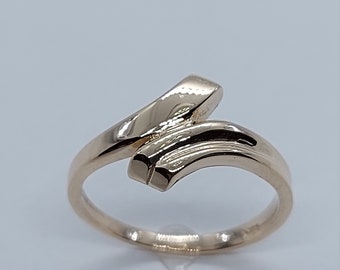 Op de afbeelding: Een ring van goudkleurig metaal met een uniek, gebogen ontwerp. De band is dun en de bovenkant van de ring heeft een gebogen, plat stuk metaal dat schuin is.