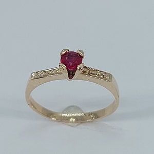 May include: A gold ring with a single red gemstone set in a four-prong setting. The ring has a thin band with small clear stones set in a pave setting.