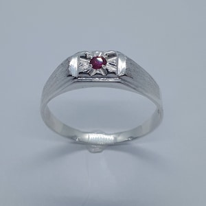 May include: A silver ring featuring a rectangular face with a small, round, red gemstone. The ring has a textured band and a decorative design around the gemstone. The ring is a classic style.