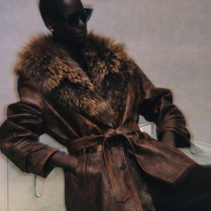 May include: A brown leather coat with a fur collar and a matching belt. The coat has a textured appearance and is buttoned at the cuffs. The fur collar is a mix of brown and tan colors.