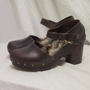 Może przedstawiać: Para brązowych skórzanych chodaków z futrzaną podszewką i paskiem z klamrą. Chodaki mają drewniany obcas i są ozdobione małymi, złotymi ćwiekami wokół podstawy. Buty mają zamknięty nosek.