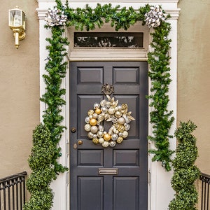 Puede incluir: Una puerta de entrada gris oscuro con una corona navideña dorada y blanca decorada con adornos. La puerta está flanqueada por dos árboles de topiario verde en urnas negras. Una guirnalda de vegetación con lazos plateados cuelga sobre la puerta.