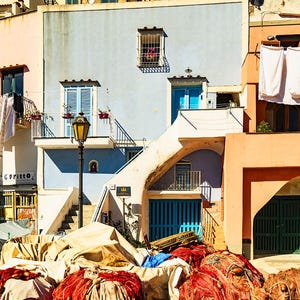 May include: A colorful building with blue, pink, and orange walls. The building has several windows and doors, and there are white sheets hanging on a clothesline. The building is located in a coastal town and there are fishing nets in the foreground.