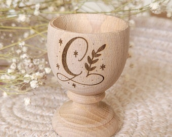 Puede incluir: Un porta huevos de madera con un diseño tallado de una letra C, una hoja y estrellas. El porta huevos es de color marrón claro y tiene una base redonda.