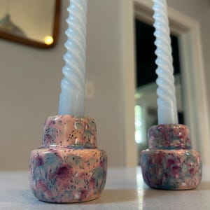 Puede incluir: Dos portavelas de cerámica con forma redondeada y cónica, con un esmalte moteado en tonos rosa, azul y morado. Cada portavelas contiene una vela larga de color blanco con forma de espiral retorcida colocada verticalmente.
