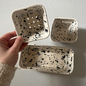 Può includere: Un set di tre contenitori in ceramica con un design maculato nero e crema. Il set comprende un contenitore quadrato con fori di drenaggio, un contenitore quadrato più piccolo e un contenitore rettangolare.