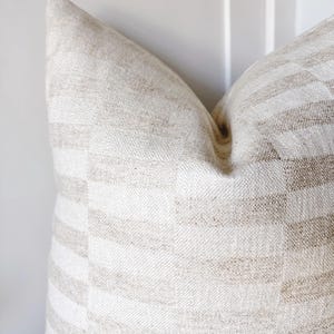 May include: A square throw cushion featuring a woven fabric with a repeating checkered pattern in alternating shades of beige and off-white. The textile has a visible, textured grain throughout, creating a subtle contrast between the light and dark squares.