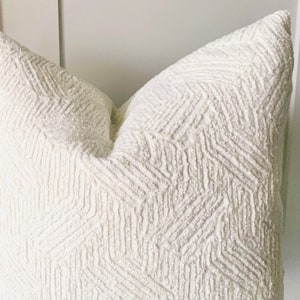 May include: A square throw cushion cover in a solid off-white colour. The fabric features a raised, textured geometric pattern consisting of repeating chevron-like lines and small, nubby tufted details across the entire surface of the textile.