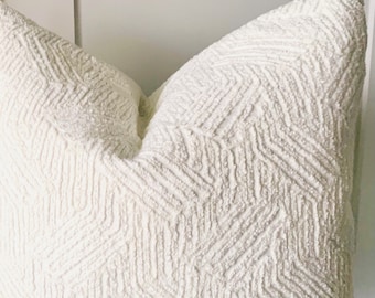 May include: A square throw cushion in a solid off-white colour featuring a raised, textured geometric pattern. The surface is covered in a repeating chevron-like design created with thick, nubby fabric threads that provide a tactile finish.