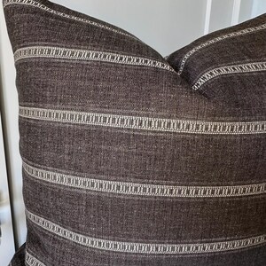 May include: A square throw pillow featuring a dark charcoal gray fabric with a visible woven texture. The pillow is decorated with horizontal stripes in a lighter cream or off-white color, which include intricate, repeating geometric patterns.