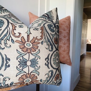 May include: Three decorative throw pillows. The front pillow has a cream background with a floral pattern in teal, brown, and blue. The second pillow is a solid peach color. The third pillow has a peach background with a repeating wave pattern.