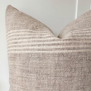 May include: A close-up of a decorative pillow with a textured, woven fabric. The pillow features horizontal stripes in a lighter shade against a darker brown background. The pillow is a neutral color, suitable for various home decor styles.