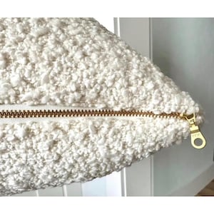 May include: A cream-colored textured bouclé pillow cover featuring a prominent gold-toned metal zipper running horizontally across the center. The fabric has a thick, nubby, and looped surface texture, and the zipper pull is clearly visible.