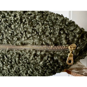 May include: A dark olive green textured sherpa fabric cushion cover featuring a gold-toned metal zipper closure. The zipper pull is clearly marked with the letters YKK, indicating the brand of the hardware on this soft, curly-textured item.