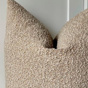 May include: Close-up of a textured, beige decorative cushion. The cushion has a nubby, woven appearance, with a neutral colour palette. The cushion's shape is a classic square, perfect for adding texture to any room.