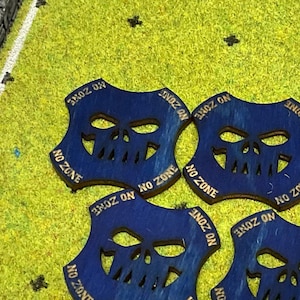 May include: Six blue wooden tokens with a skull design and the text "NO ZONE" printed on each. The tokens are arranged in a circle on a green surface.