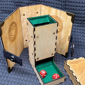 Puede incluir: Un juego de torre de dados y bandeja de juego de madera con interior de fieltro verde. La torre es rectangular con detalles cortados con láser, y la bandeja contiene dos dados rojos. Una pantalla de juego de madera está detrás de la torre.