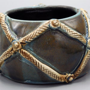 May include: A small, round ceramic bowl with a dark brown glaze and a light brown, woven design. The bowl has a unique, textured surface.