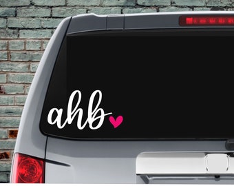 May include: A white vinyl decal with the text "ahb" and a pink heart on a black car window.