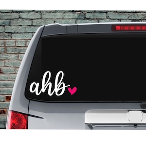 May include: A white vinyl decal with the text "ahb" and a pink heart on a black car window.