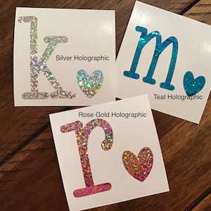 May include: Three vinyl stickers with glitter letters and hearts. The letters are K, M, and R. The hearts are silver, teal, and rose gold. The text below each sticker says "Silver Holographic", "Teal Holographic", and "Rose Gold Holographic".