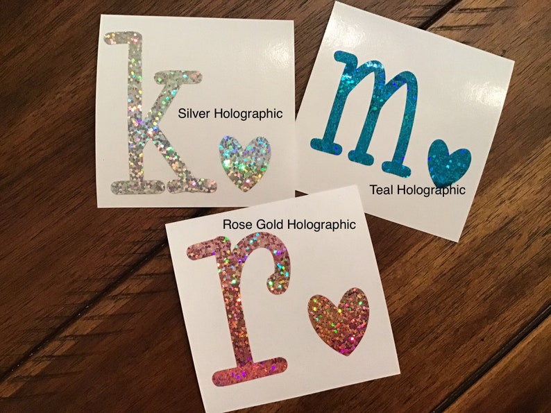 May include: Three vinyl stickers with glitter letters and hearts. The letters are K, M, and R. The hearts are silver, teal, and rose gold. The text below each sticker says "Silver Holographic", "Teal Holographic", and "Rose Gold Holographic".