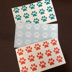 May include: Three sheets of white paper with paw prints in different colors. The top sheet has teal paw prints, the middle sheet has white paw prints, and the bottom sheet has orange paw prints.
