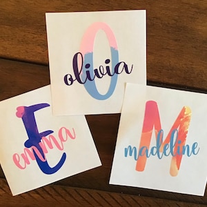May include: Three personalized stickers with names "Olivia", "Emma", and "Madeline" in a colorful watercolor style. The letters are outlined in blue and filled with pink and orange gradients.